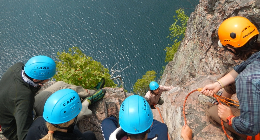 Four people wearing safety gear peer over the edge of a cliff, looking down at a rock climber making their way up. They are all above a blue body of water.